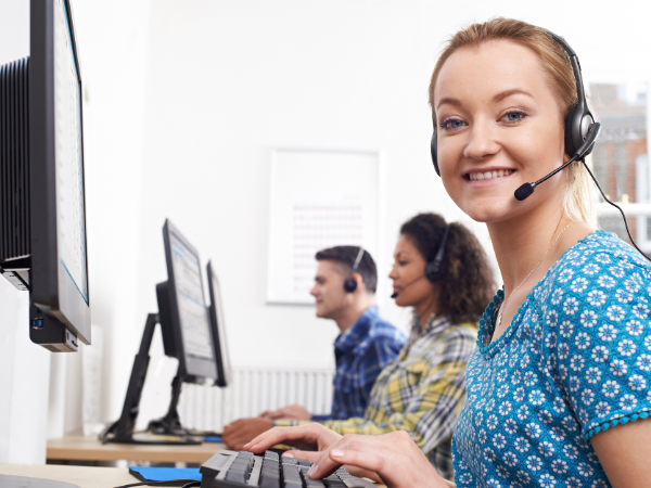 A smiling woman in a blue shirt wearing a headset works at a computer in a customer service call center, representing Integrity Solutions Australia's focus on customer service training. Two colleagues are in the background, also engaged in customer support tasks.